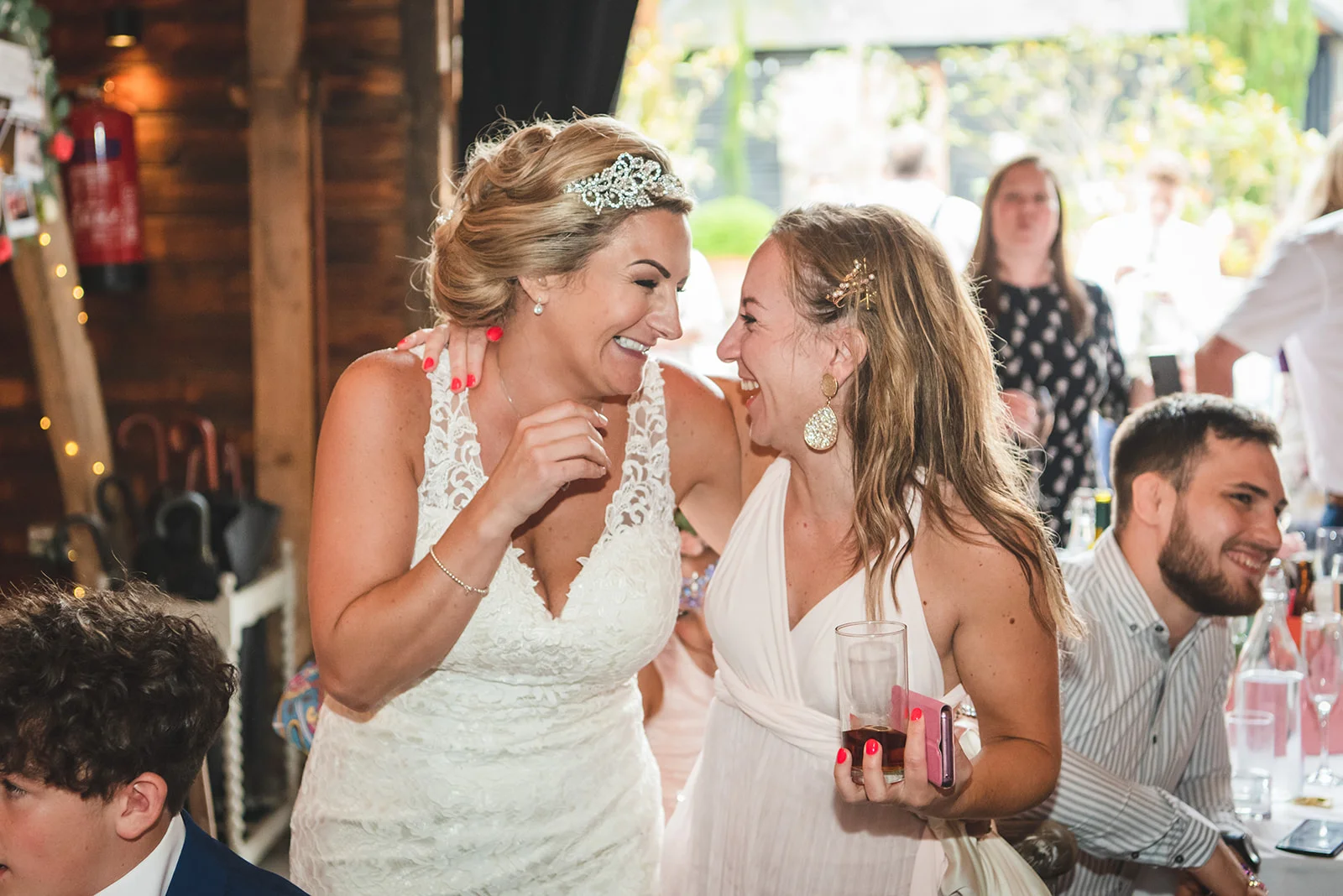 Two smiling brides share a joyful, laughing moment together during a rustic barn wedding reception, surrounded by wedding guests.