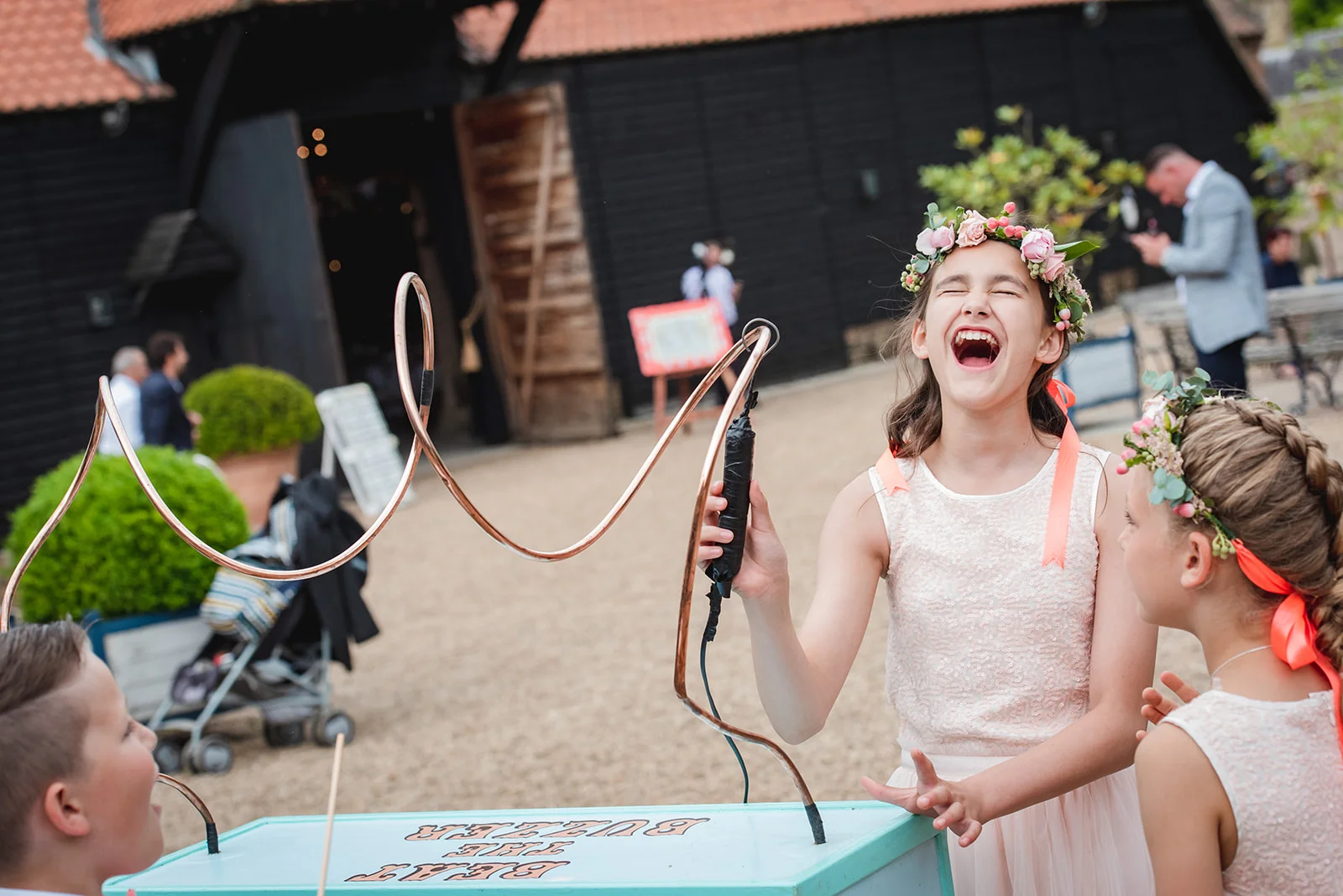 A young flower girl laughing joyfully while playing a buzzer wire game outdoors at a wedding reception near a black barn.