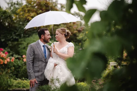 A newlywed couple shares a tender glance while walking outdoors beneath a white umbrella in a lush garden setting.