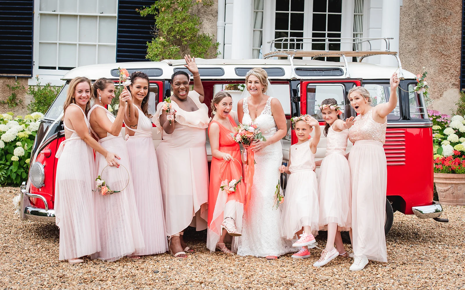 A joyful bride and her diverse bridal party in pink dresses pose playfully in front of a vintage red and white VW campervan outside a country house.