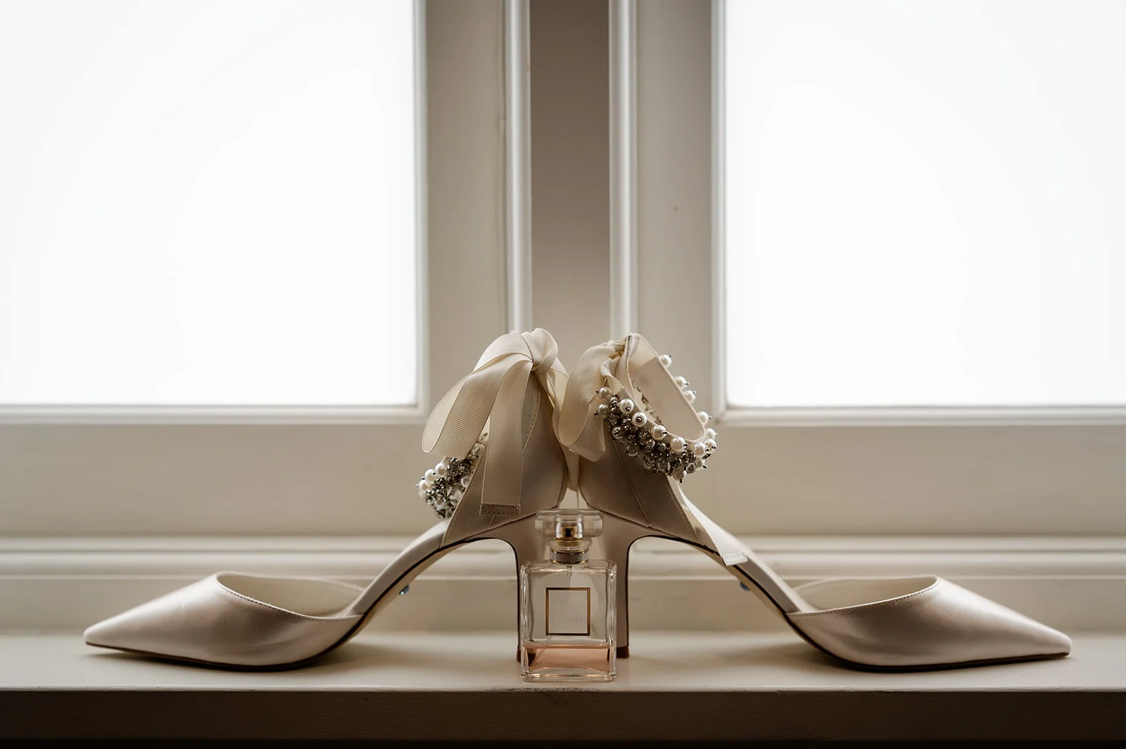 A pair of elegant satin bridal shoes with pearl and crystal ankle straps rest symmetrically on a windowsill alongside a bottle of perfume, illuminated by soft window light.