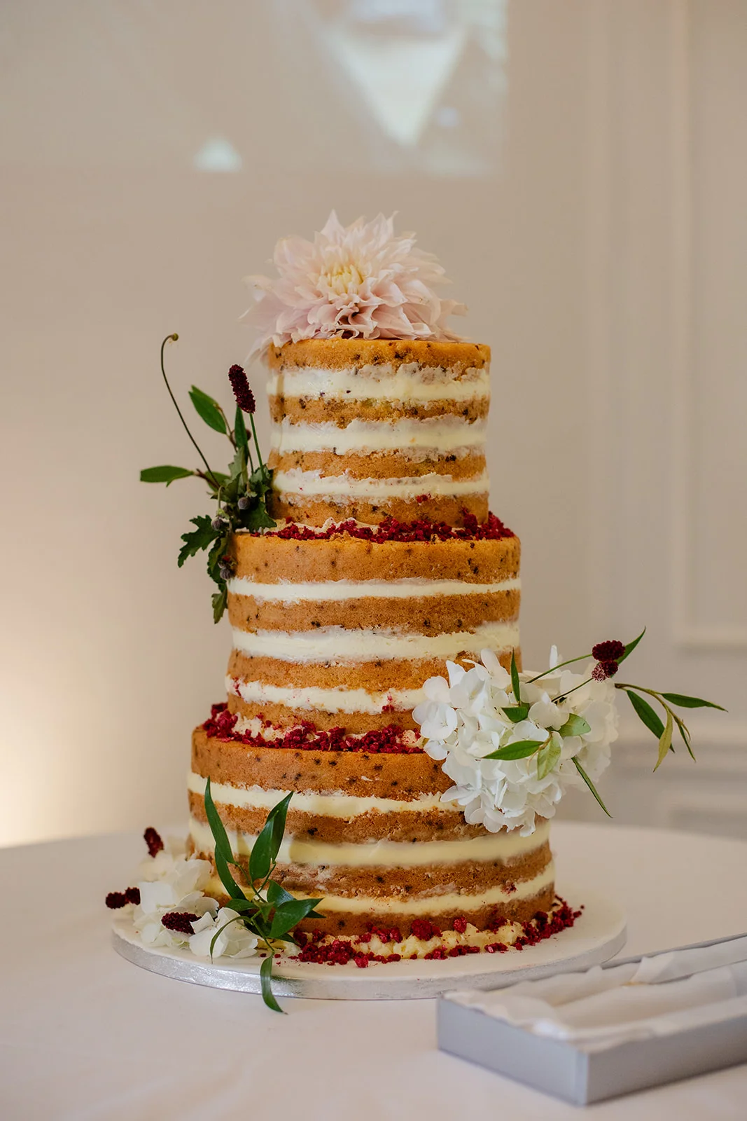 A close-up shot of a three-tier semi-naked wedding cake adorned with fresh white hydrangeas and a soft pink dahlia on top.