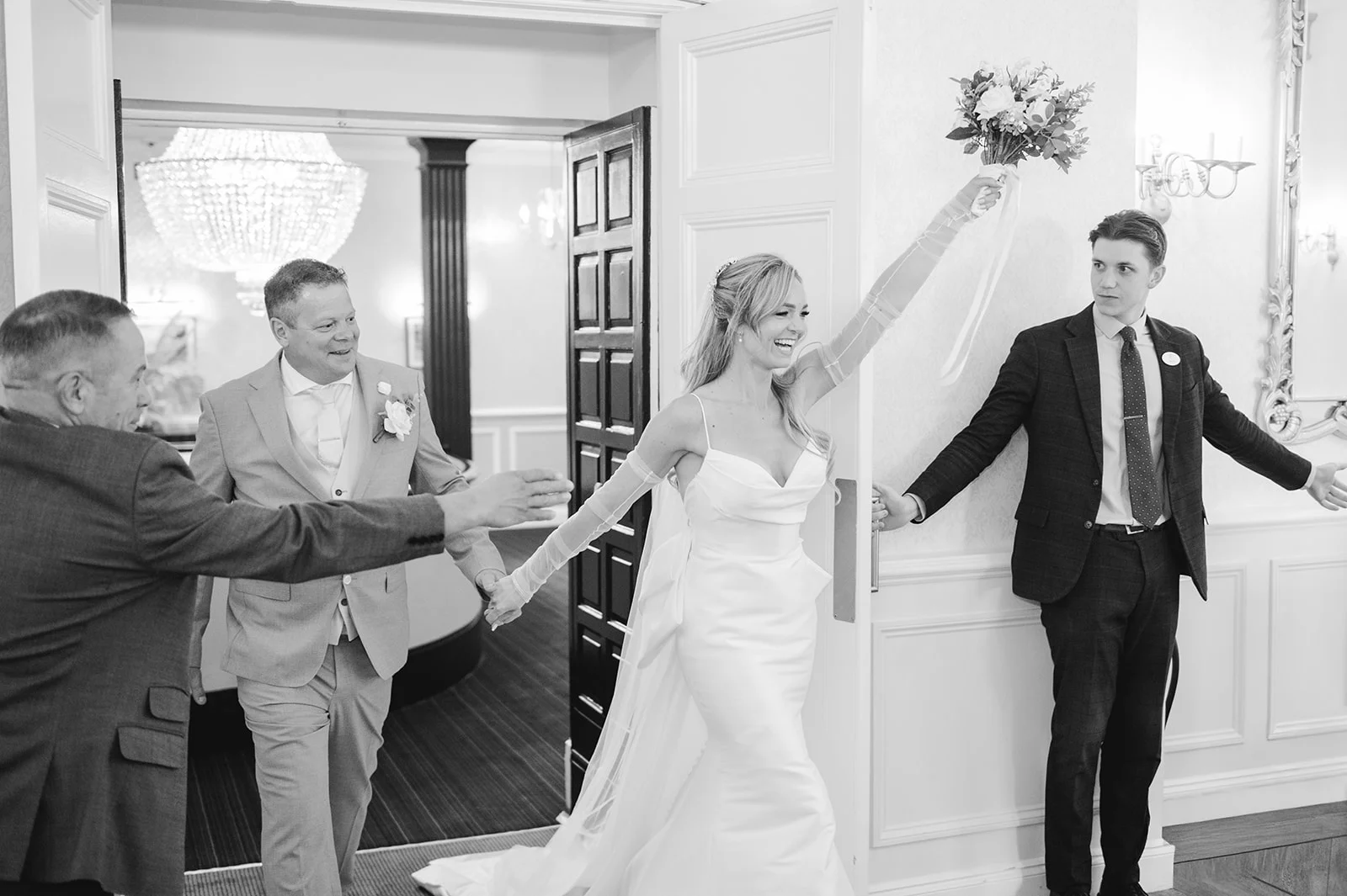 A joyful newlywed bride and groom enter their wedding reception holding hands, with the bride laughing and holding her bouquet high in the air.