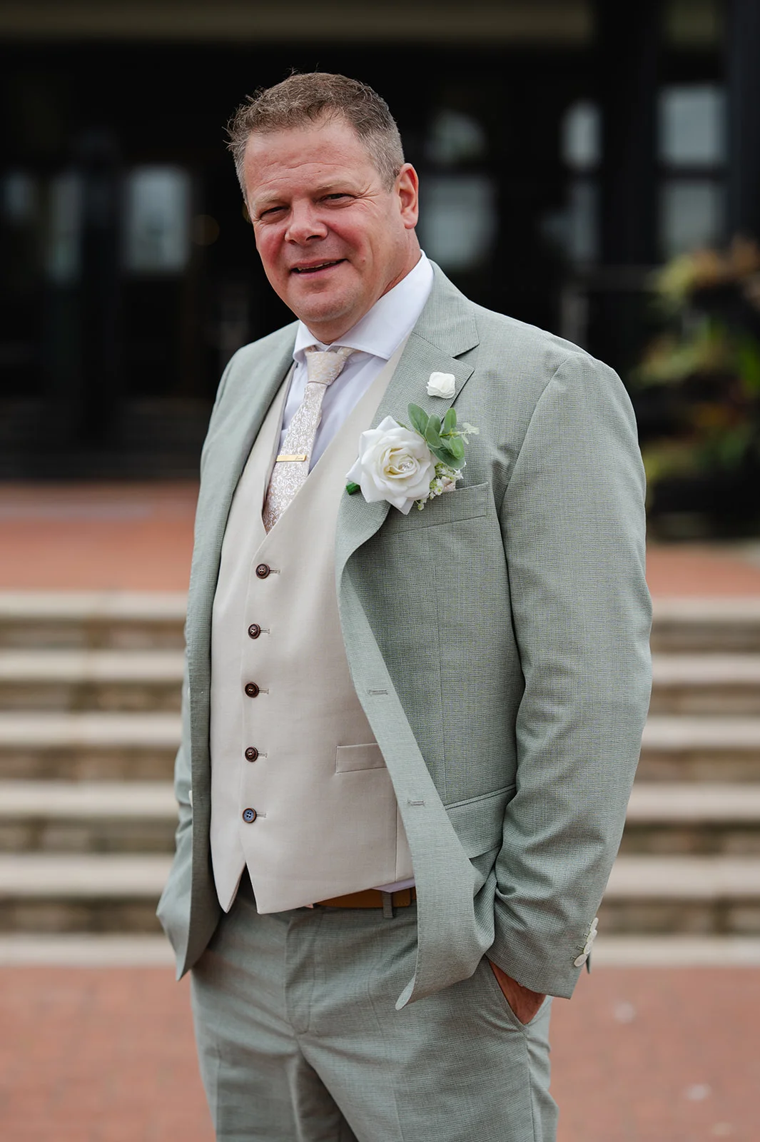 A mature groom smiling while wearing a sage green suit with a beige waistcoat and a white rose boutonniere.