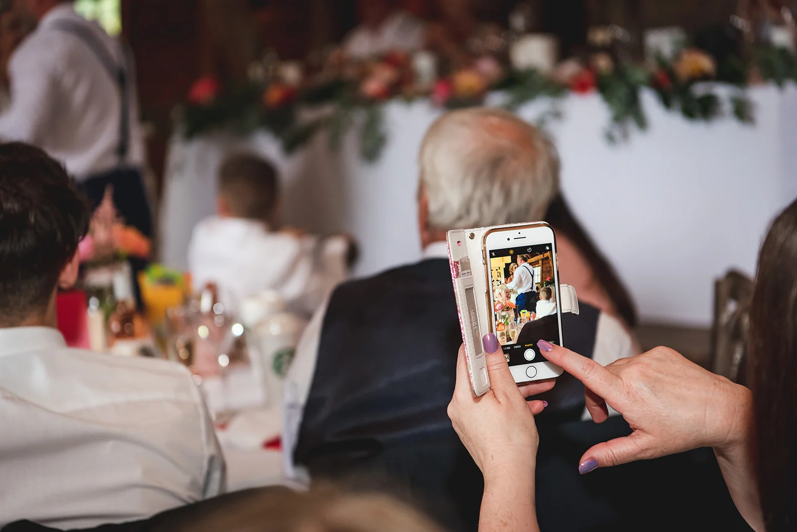 A wedding guest holds up a smartphone to photograph the speeches at the top table during the wedding breakfast.