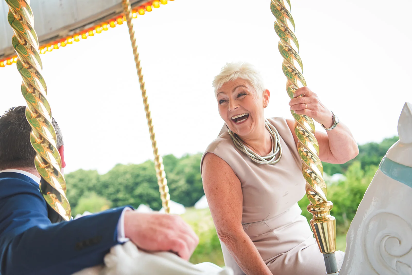 A mature bride laughs joyfully while riding a carousel at a festival-themed wedding, holding onto the polished golden pole.