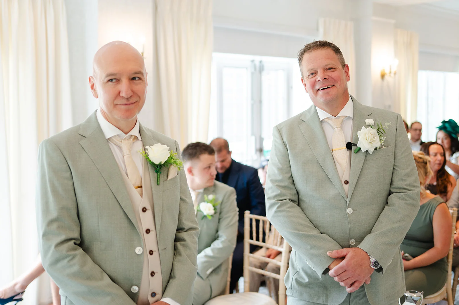 Two smiling grooms in matching sage green suits and cream ties stand side by side during their wedding ceremony inside a bright venue.