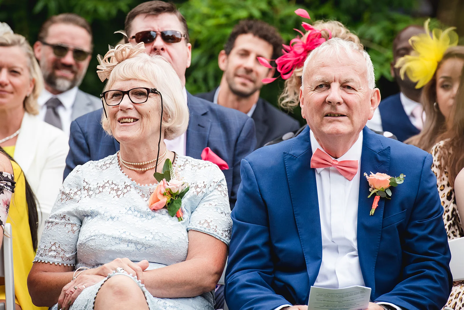 An older couple smiling happily among seated guests at an outdoor wedding ceremony, with the man wearing a smart blue suit and pink bow tie.