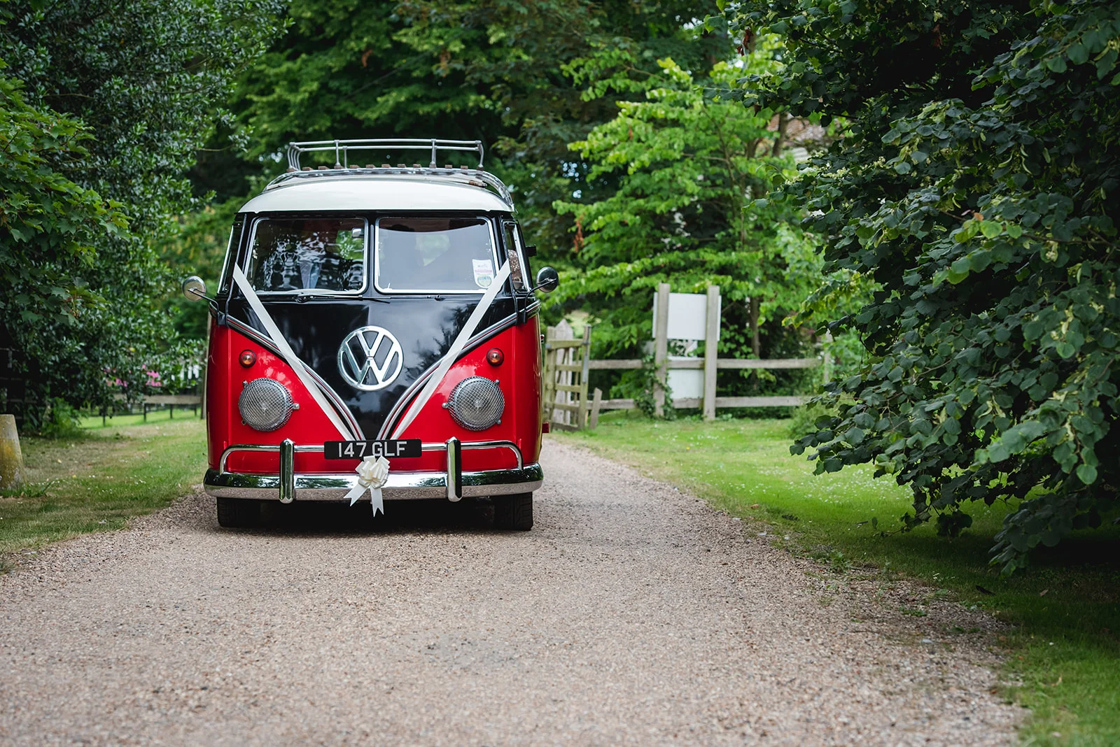 A vintage red and white Volkswagen campervan decorated with white ribbons sits parked on a gravel driveway surrounded by lush green trees.