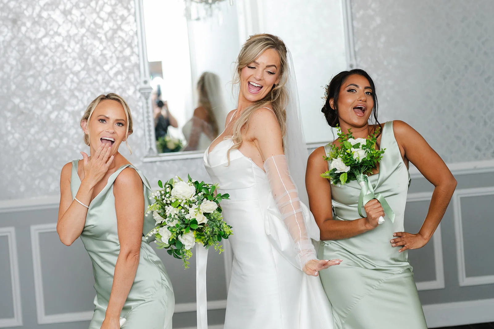 A joyful bride in a satin gown and sheer gloves laughs playfully alongside two bridesmaids in sage green dresses holding bouquets.