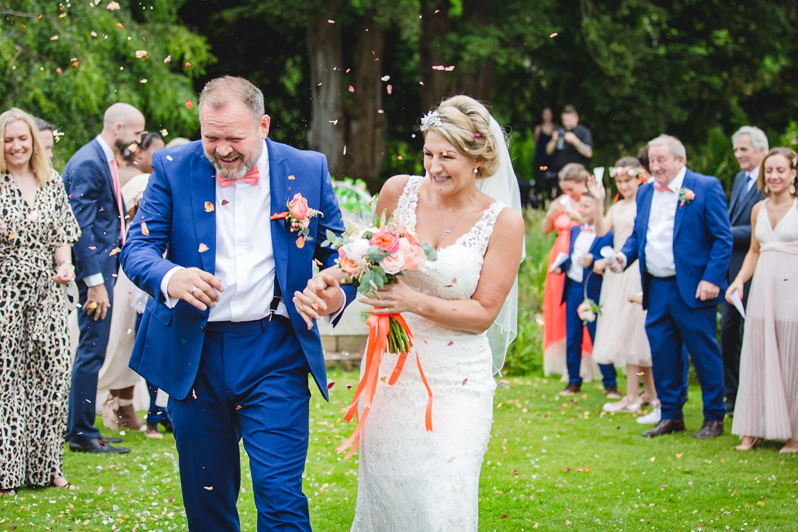 A bride and groom walk hand in hand down the lawn as guests shower them with confetti, laughing joyfully amid an outdoor garden reception.