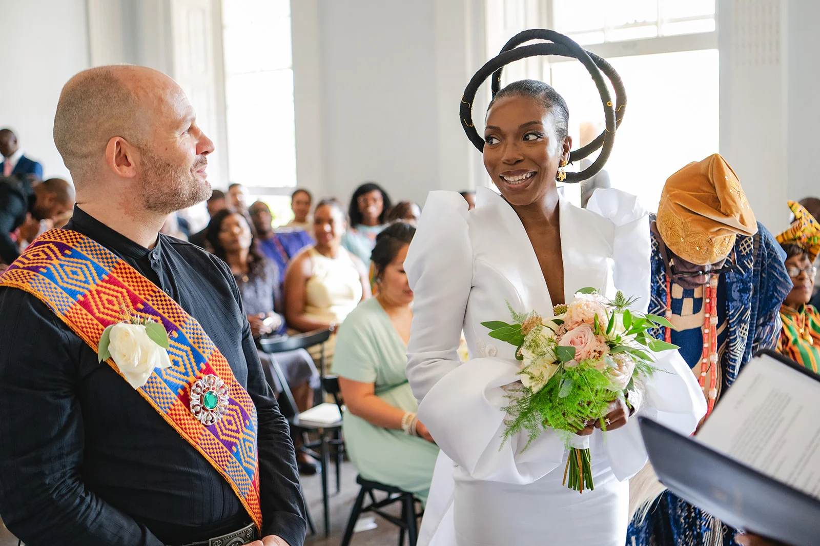 A smiling black bride wearing a structured white dress with statement sculptural headpiece and puff sleeves looks at her groom during a multicultural wedding ceremony.