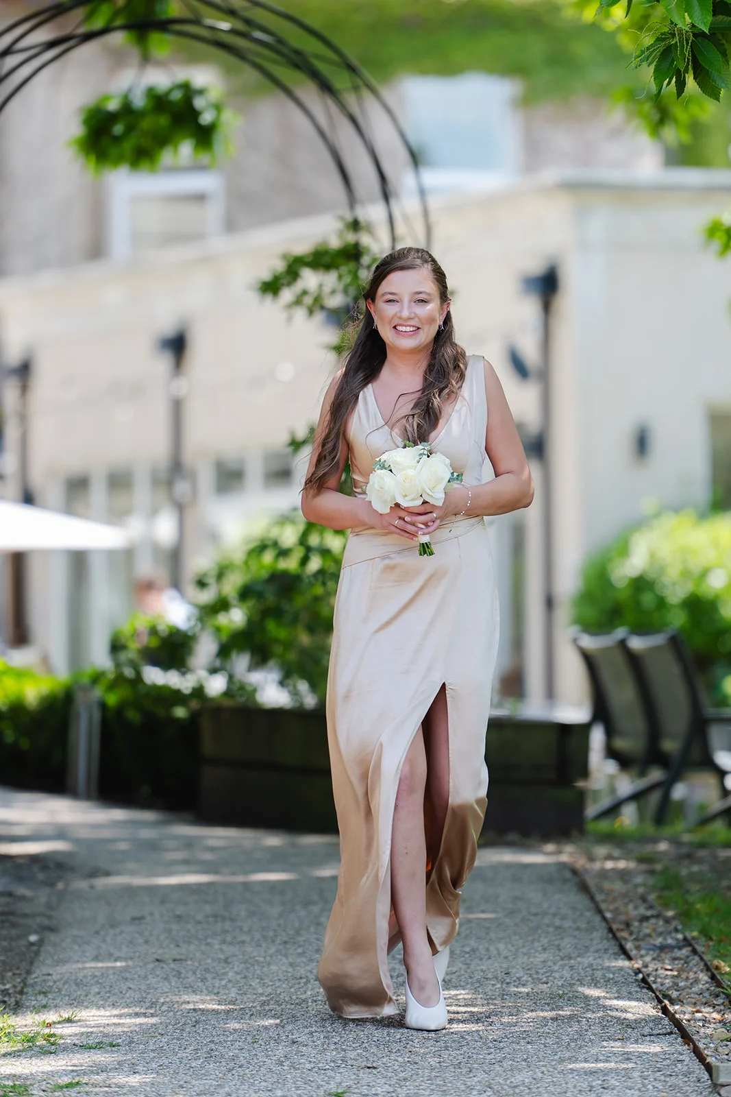 A smiling bridesmaid in a satin champagne gown with a high split walks down an outdoor pathway carrying a bouquet of white roses.