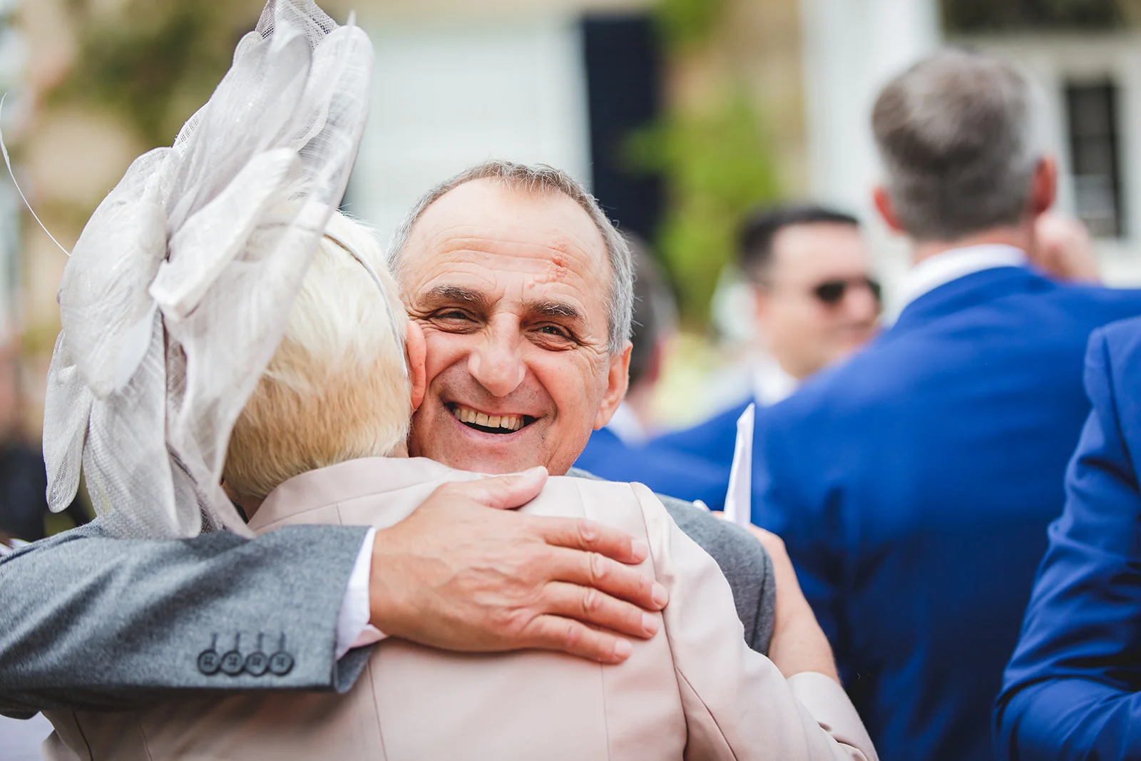 A smiling man in a grey suit hugs a guest wearing a large fascinator during an outdoor wedding reception.