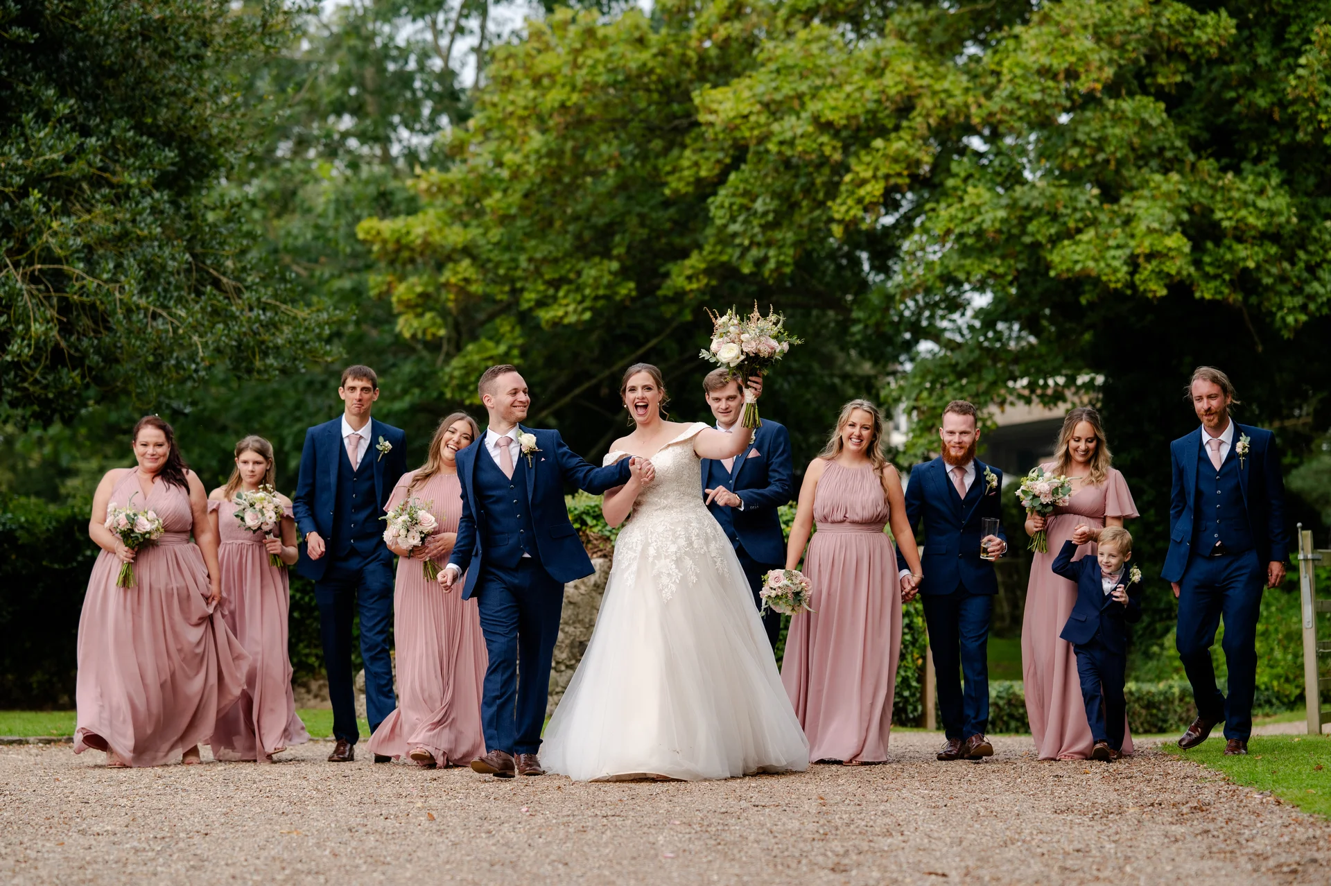 The bride and groom laugh happily as they lead their wedding party along a gravel pathway lined with lush green trees, with the bridesmaids wearing pink gowns and the groomsmen in navy suits.
