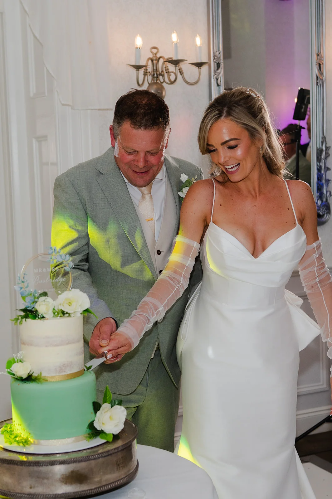 A smiling bride and groom cut their two-tier wedding cake decorated with green icing and white florals during the reception.
