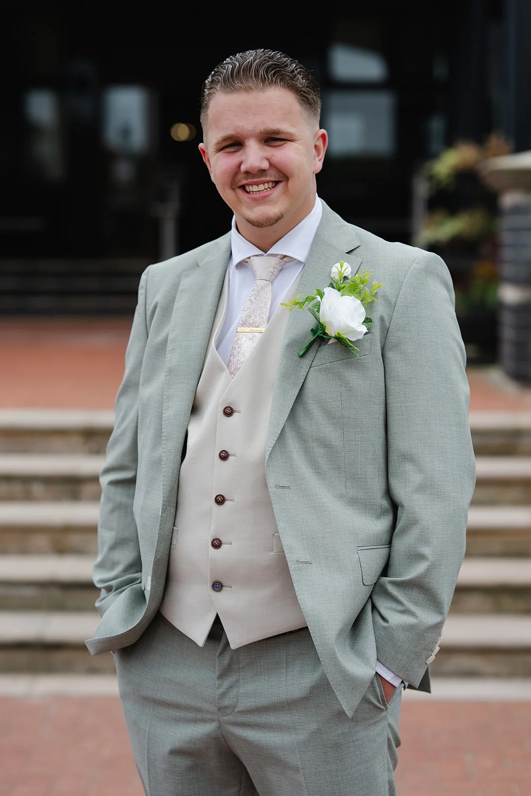 A smiling groom wearing a sage green suit, cream waistcoat, patterned tie, and a white rose buttonhole stands outdoors with his hands in his pockets.