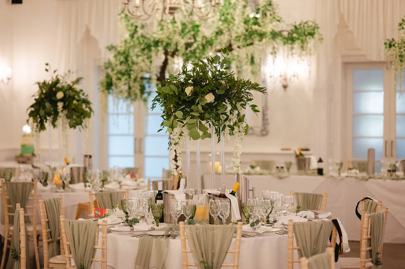 An elegant wedding reception room features round tables adorned with sage green linens, tall floral centrepieces with white roses and greenery, and a dramatic hanging floral installation overhead.