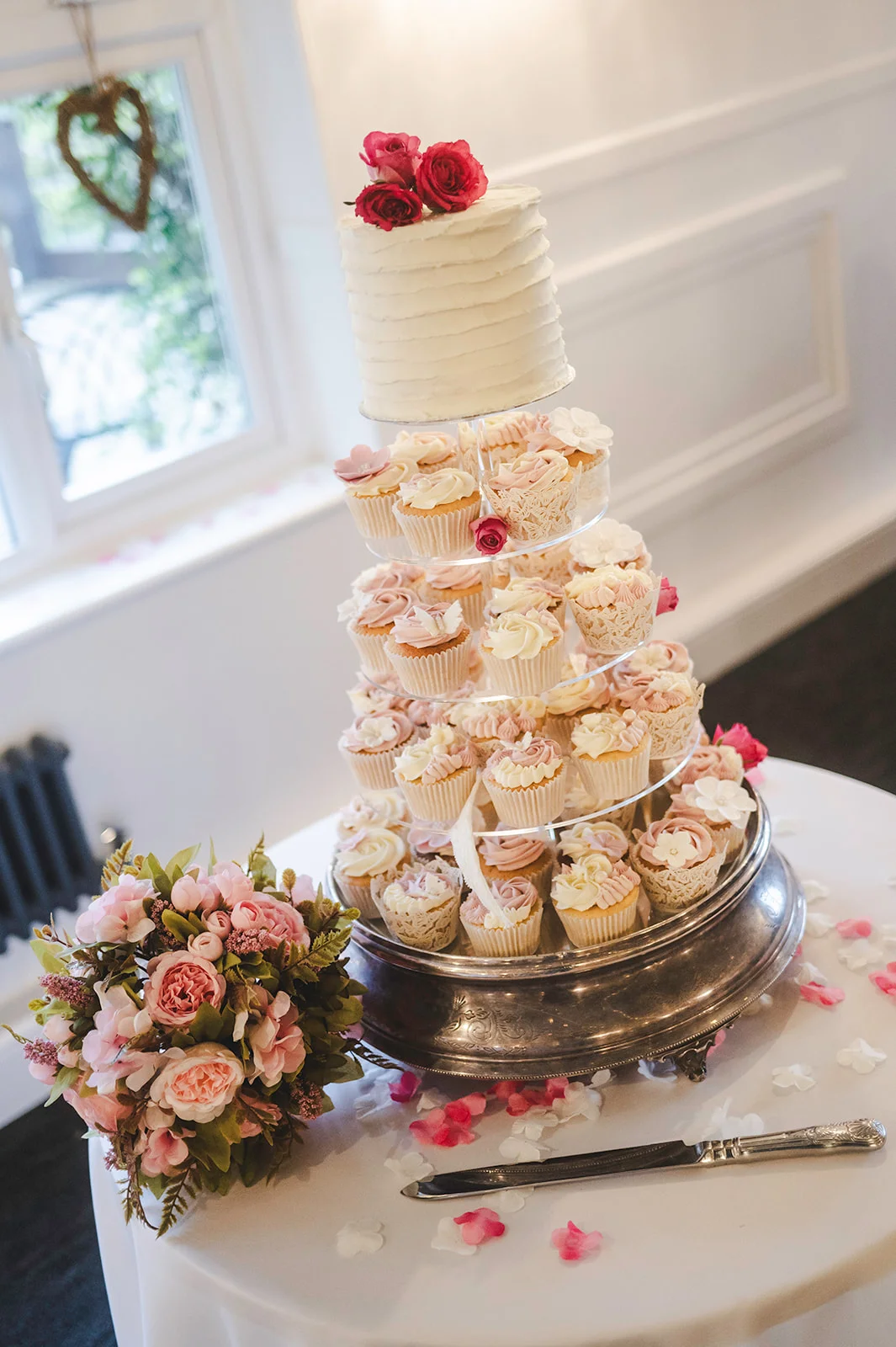 An elegant tiered cupcake tower topped with a small buttercream cake and fresh pink roses sits on a silver cake stand beside a matching floral bouquet.