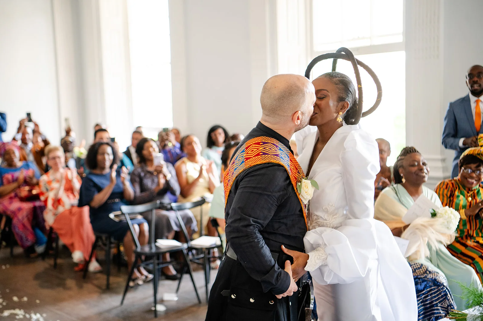 An interracial couple share their first kiss as a married couple during a bright indoor ceremony while guests applaud in the background.