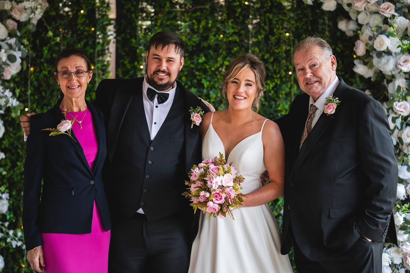 A smiling bride holding a pink bouquet stands alongside her groom in a black tuxedo and her parents against a lush green and floral backdrop.