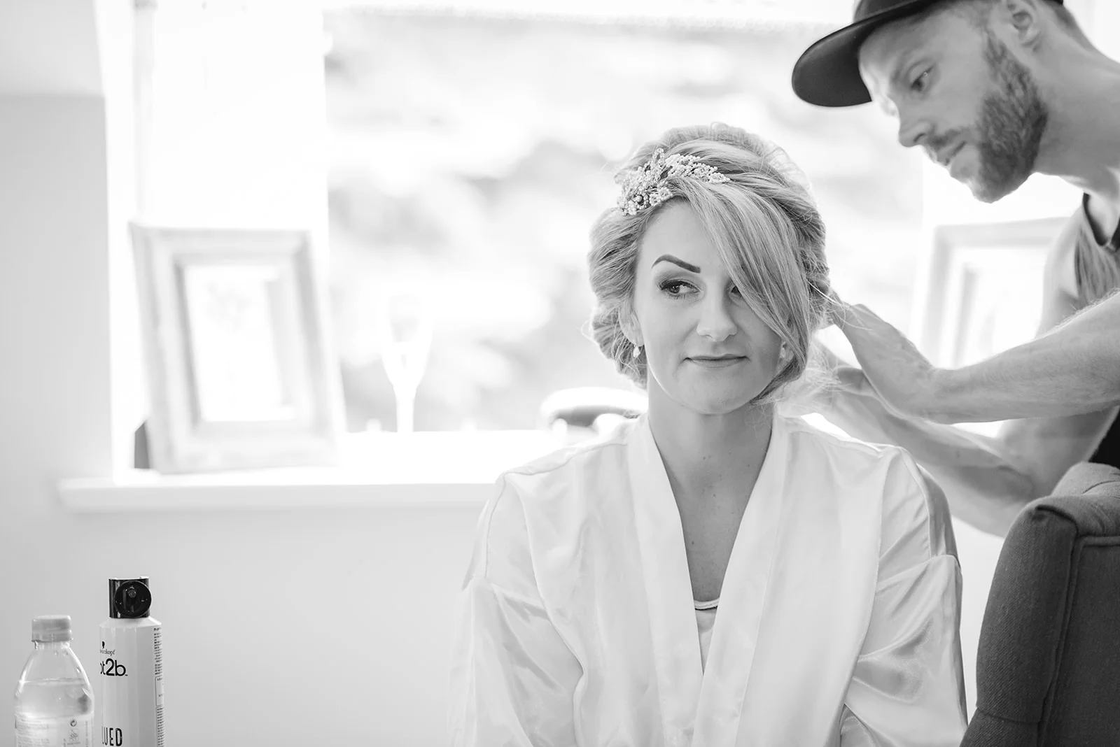 A black and white photograph captures a bride wearing a white robe and hair accessory while a stylist styles her hair indoors.