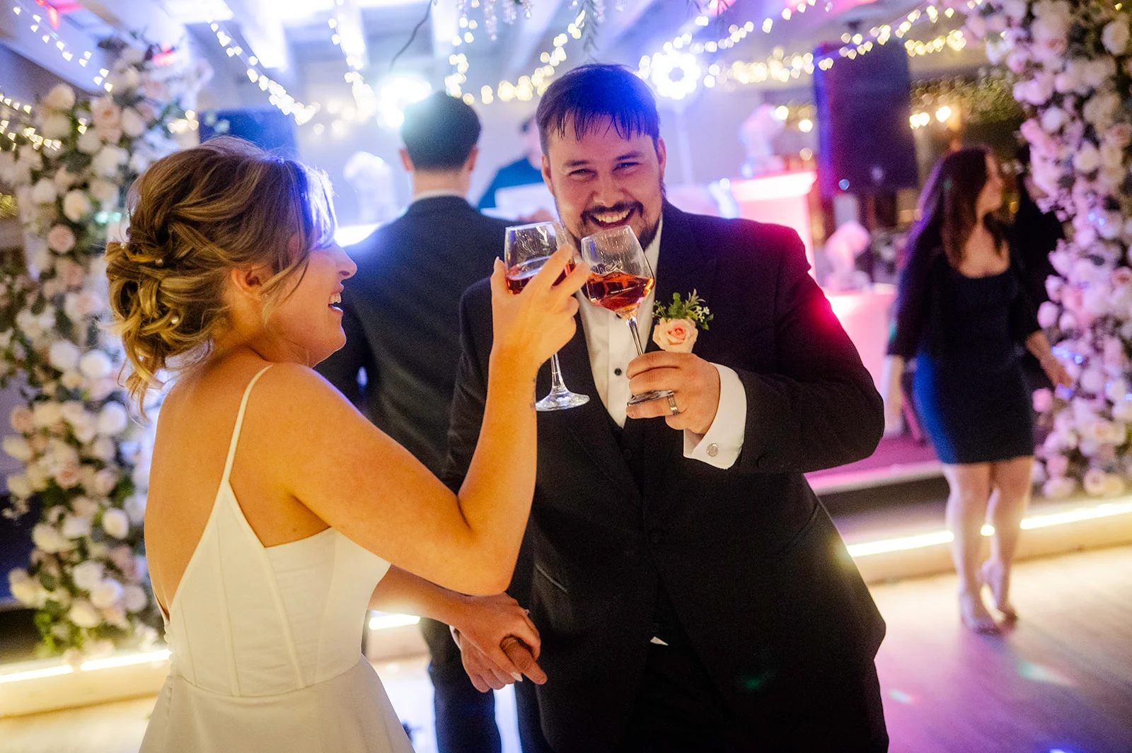 A smiling bride and groom joyfully toast each other with drinks on a brightly lit dance floor surrounded by celebratory guests and floral arches.