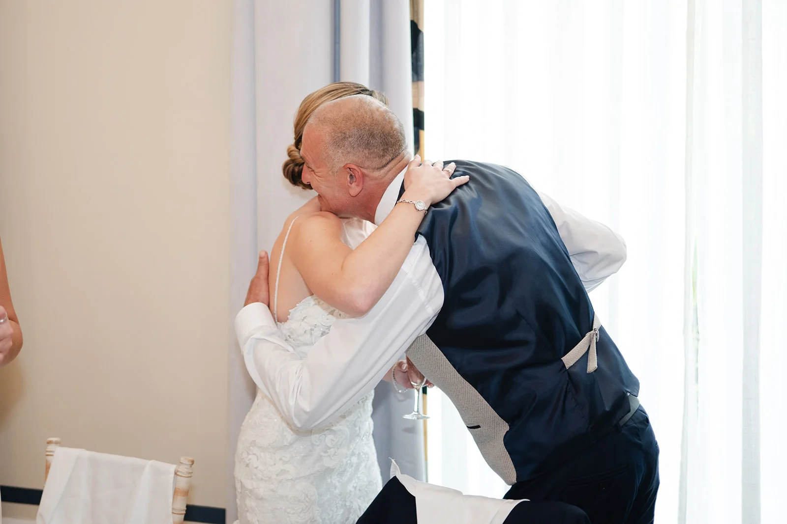 A bride in a lace wedding dress shares an emotional hug with her father during the wedding reception near a bright window.