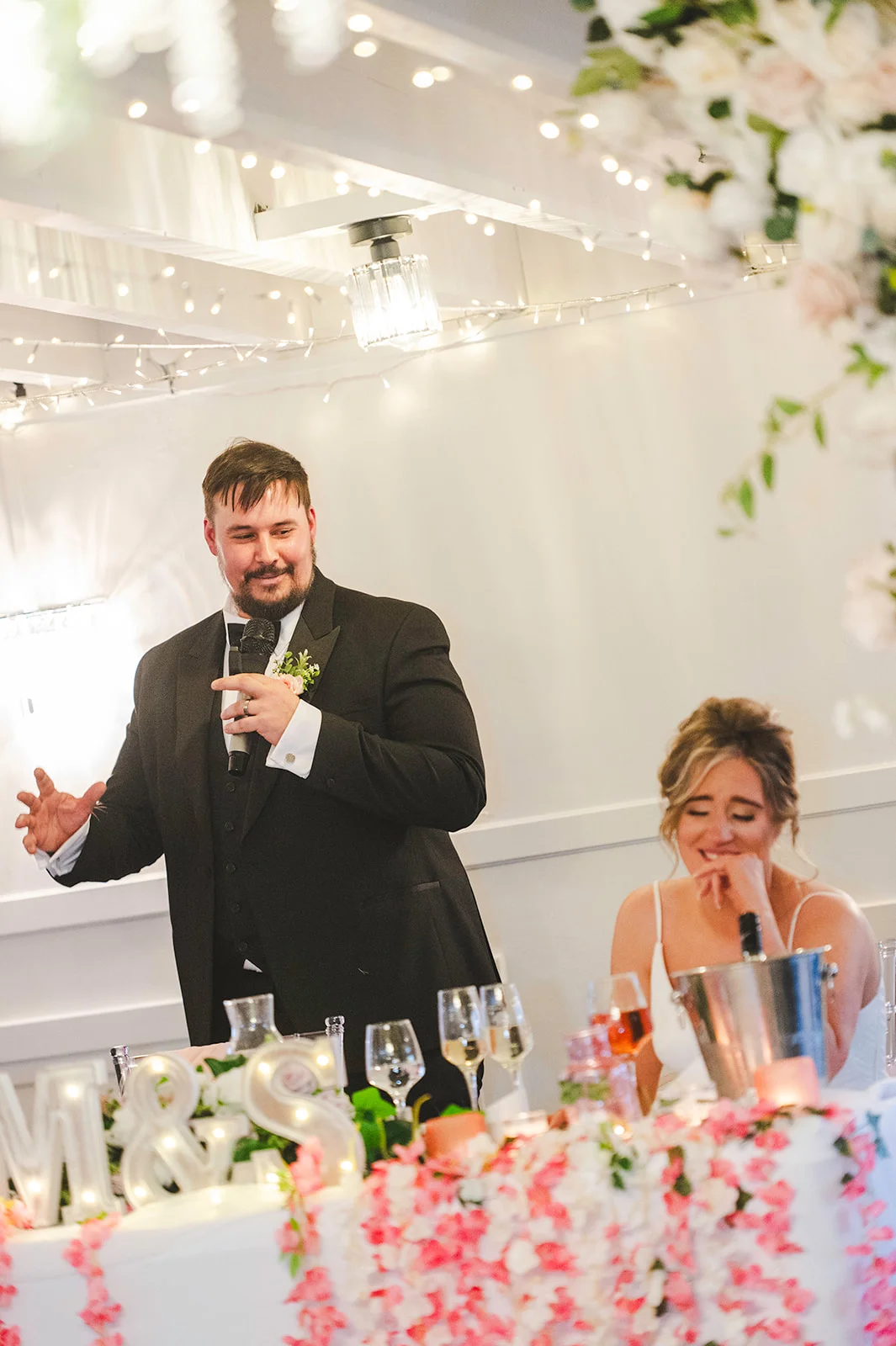 A groom delivering a speech in a black tuxedo while the bride laughs joyfully at the top table decorated with pink floral arrangements and fairy lights.