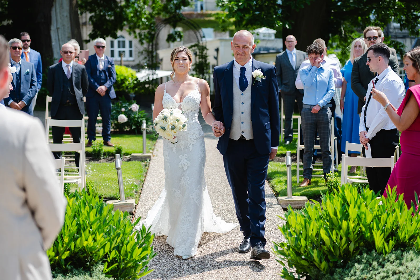 A smiling bride holding a white rose bouquet walks down the gravel garden aisle arm-in-arm with her father during an outdoor wedding ceremony.