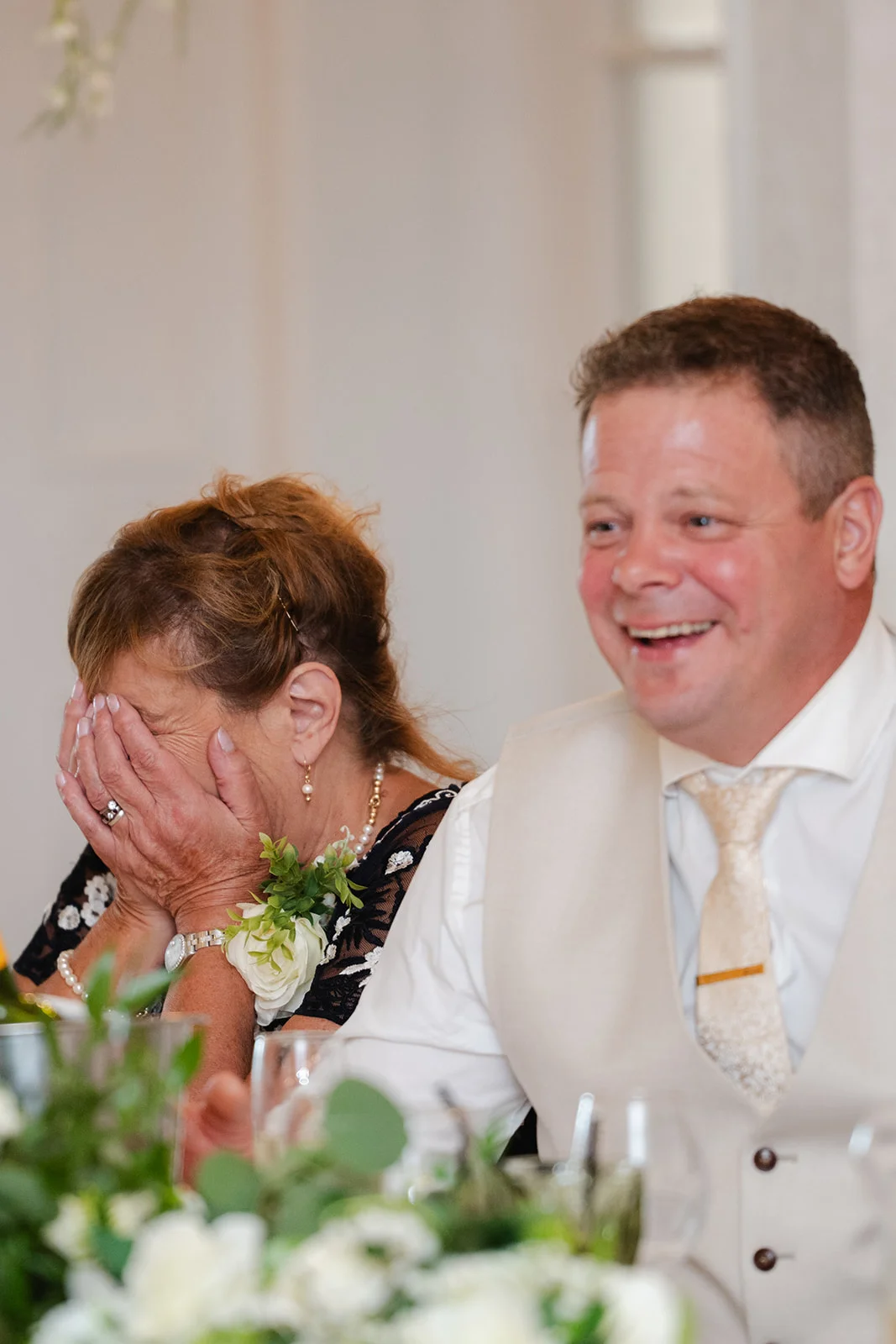 A wedding guest emotionally covers her face in tears of laughter while seated next to a smiling man during the wedding speeches.