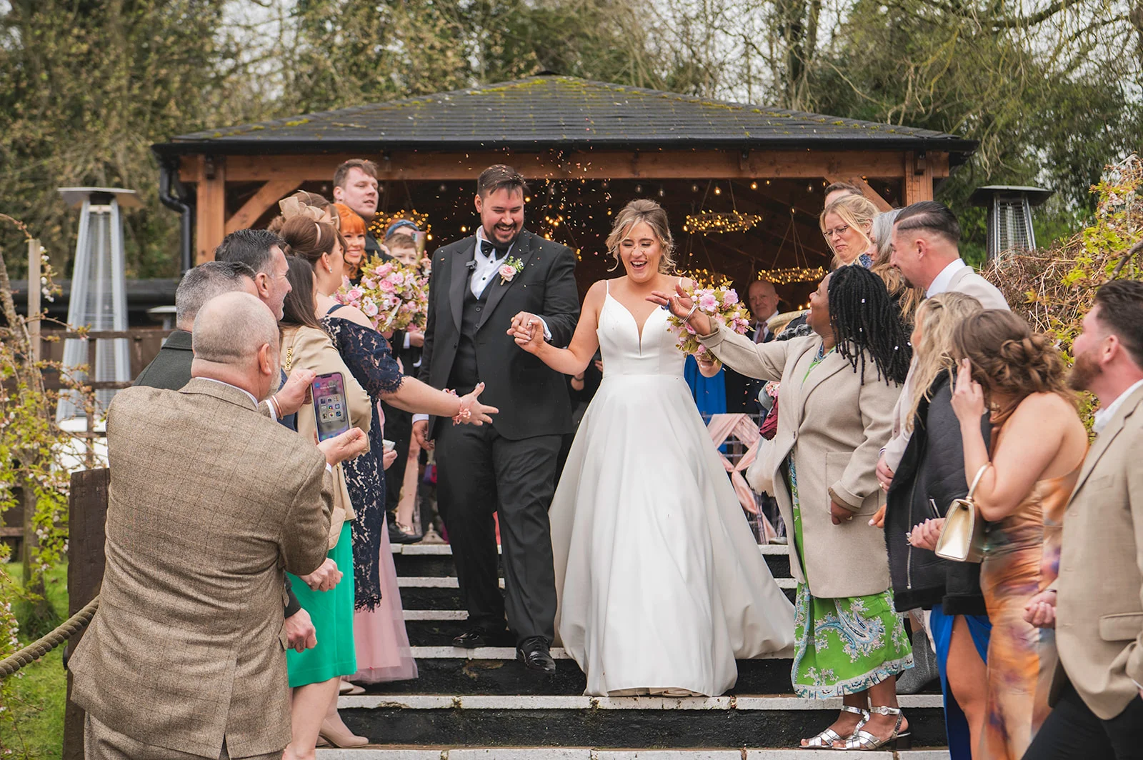 The joyful newlyweds walk down outdoor steps hand in hand as cheering guests throw confetti over them outside a wooden gazebo.