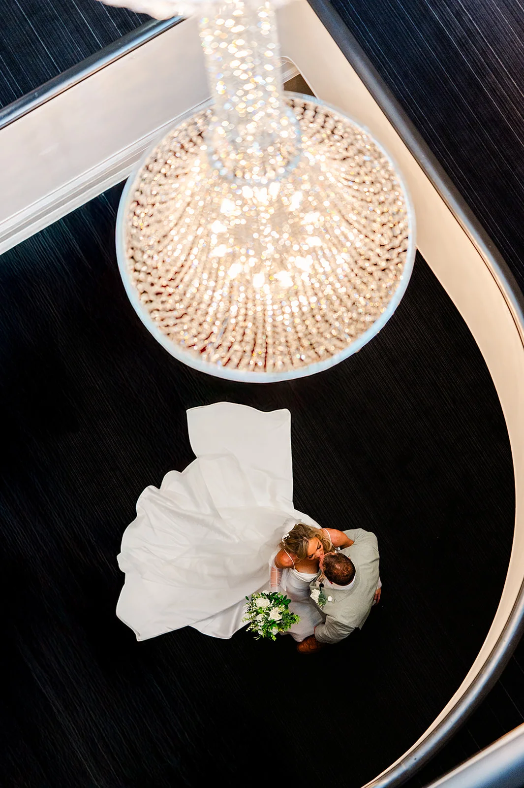 An overhead view of a newlywed couple kissing below a magnificent crystal chandelier on a sweeping spiral staircase.