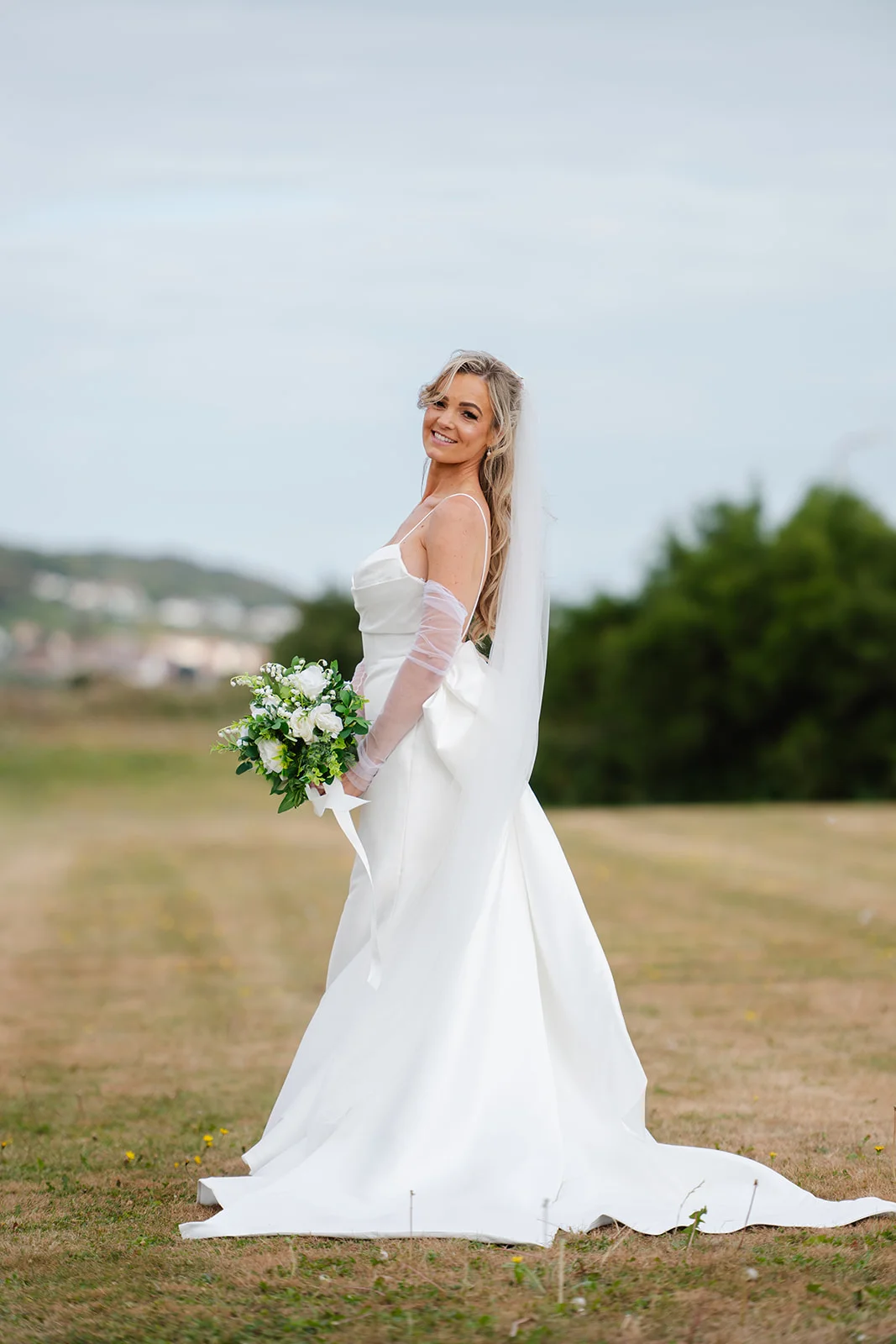A smiling bride in an A-line satin gown with sheer gloves and a chapel veil stands outdoors holding a white rose bouquet.