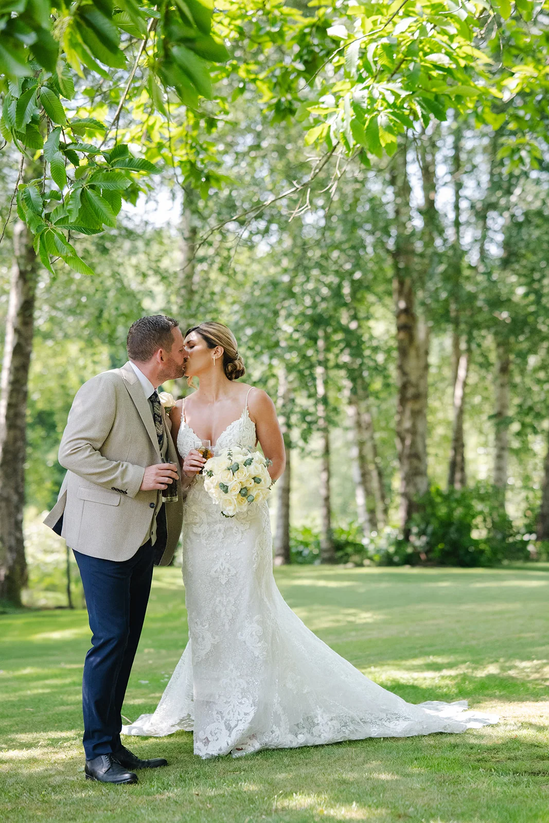 Newlyweds share a tender kiss in a sunlit woodland clearing surrounded by lush greenery and towering trees.