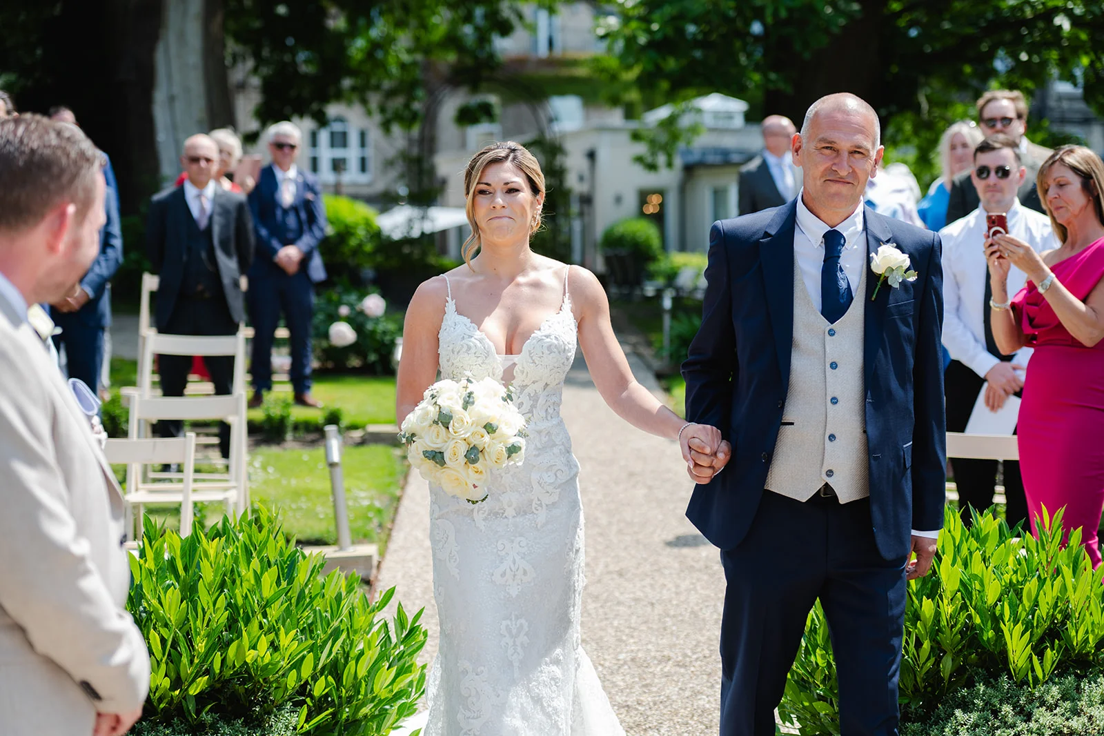 The emotional bride walks down the outdoor aisle holding hands with her father, carrying a classic white rose bouquet towards the groom.