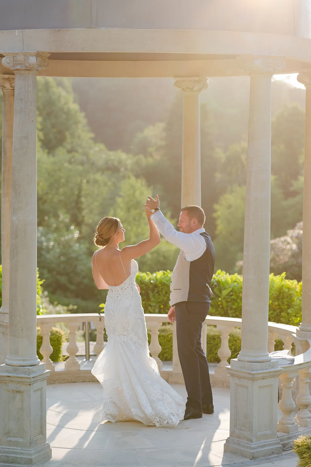 A newlywed couple shares a dance inside a stone gazebo bathed in warm golden hour sunlight during their outdoor wedding portrait session.