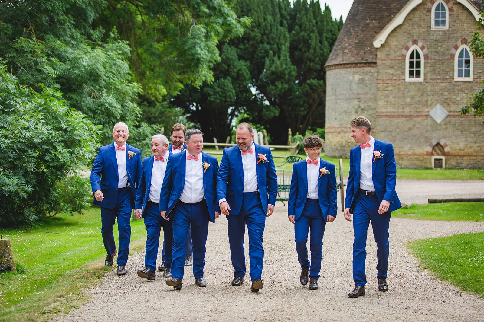 A group of groomsmen in matching blue suits and pink bowties walk joyfully along a gravel path next to a brick building and lush green trees.