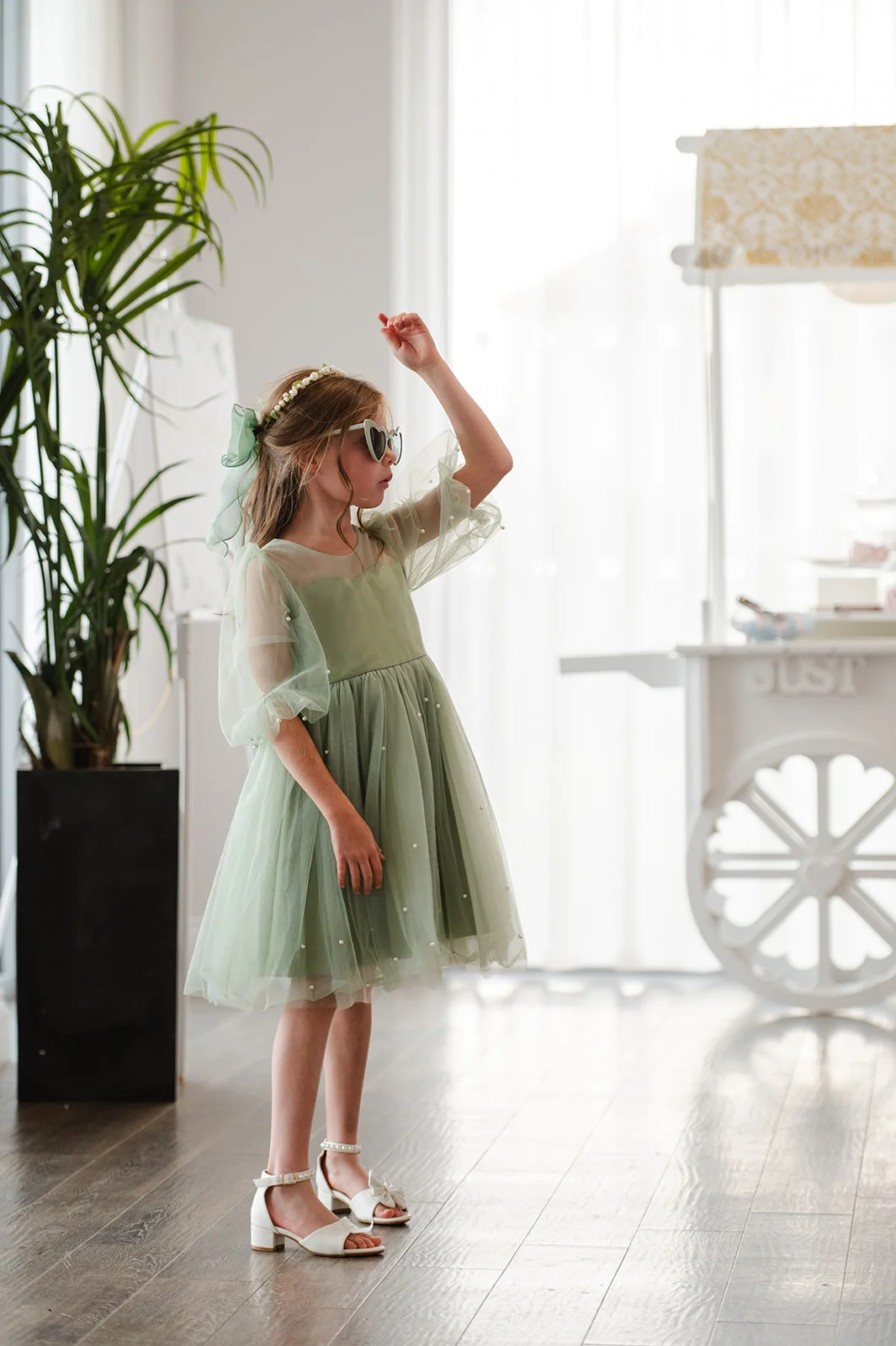 A young flower girl wearing a sage green tulle dress and stylish sunglasses poses playfully indoors near a potted plant.