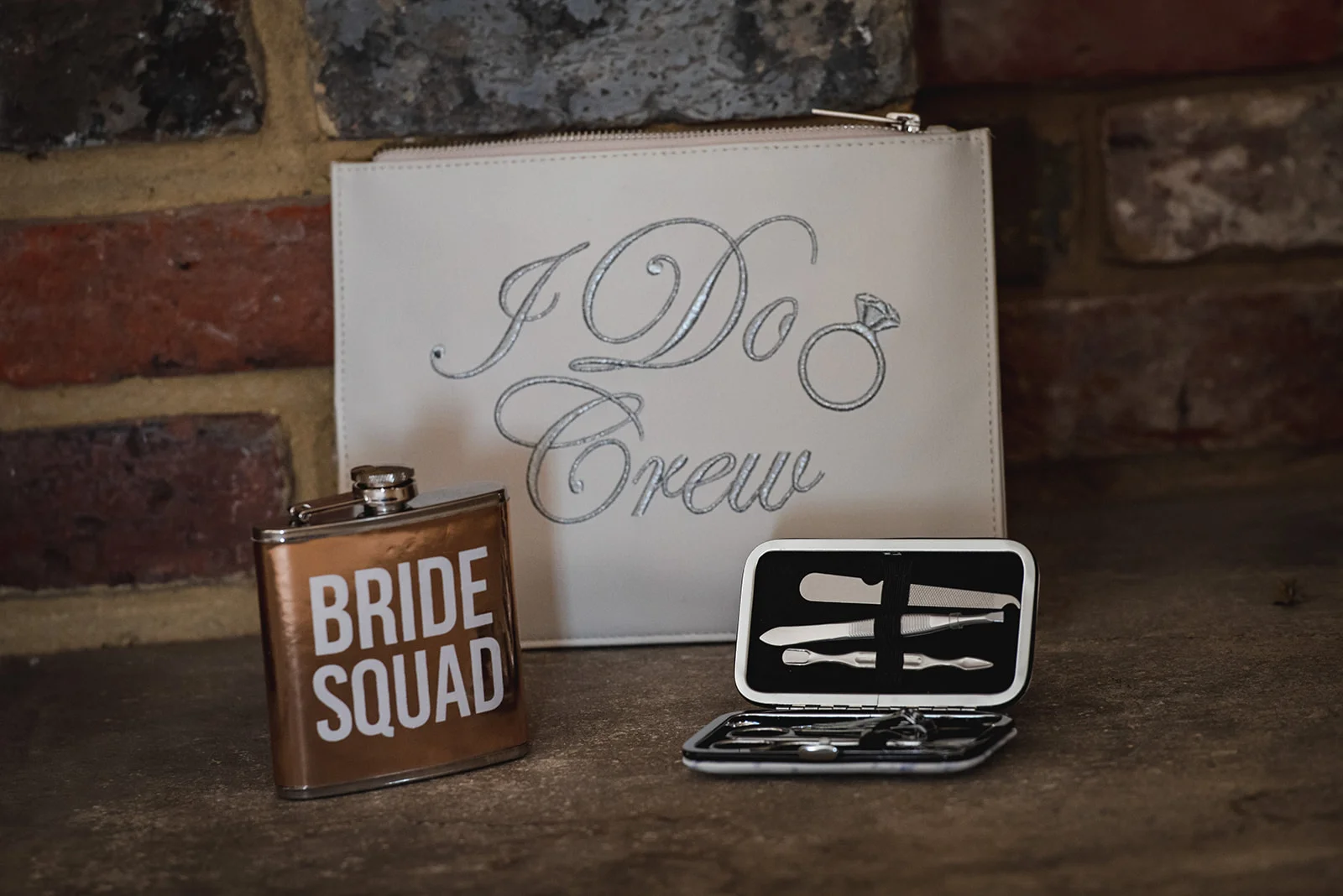 A flat lay of bridal morning accessories including a Bride Squad flask, an I Do Crew pouch, and a manicure set against an exposed brick wall.