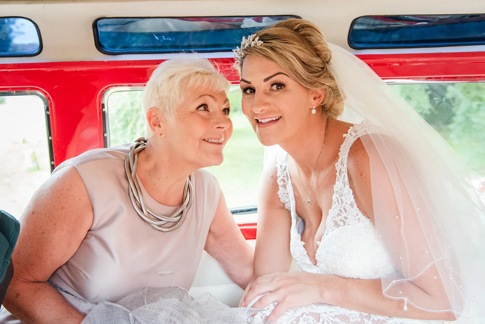 A joyful bride in a lace wedding dress and tiara shares a tender moment with her mother inside a classic VW campervan.