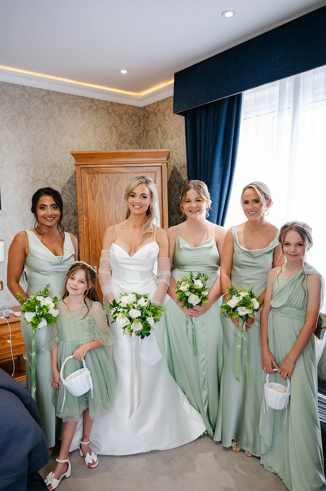 A smiling bride in a modern satin gown with sheer arm sleeves poses alongside her bridesmaids and two flower girls in sage green dresses holding white floral bouquets.