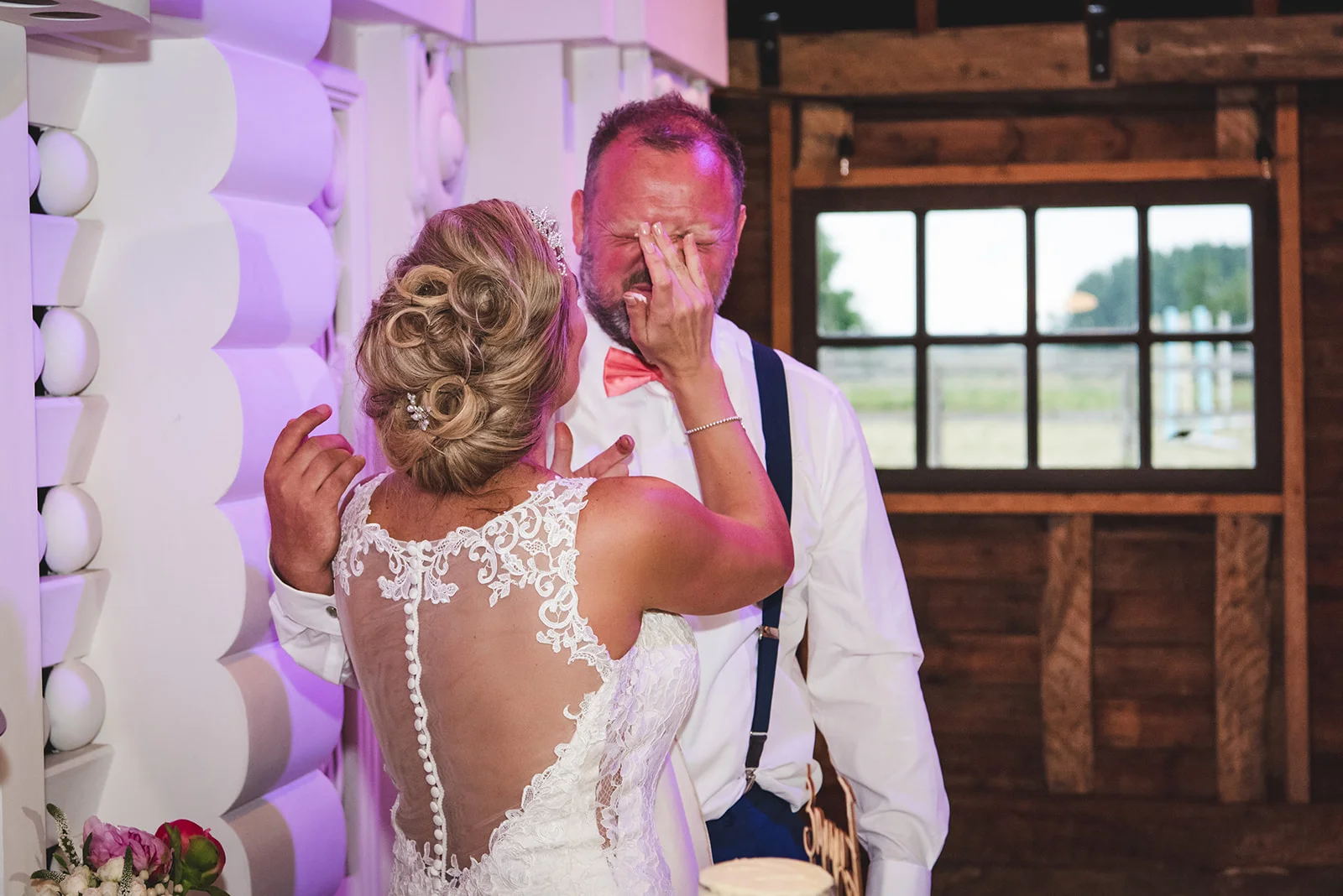 An emotional groom wipes away happy tears while standing next to his bride, who wears a lace wedding dress with a button-detail illusion back inside a rustic barn venue.