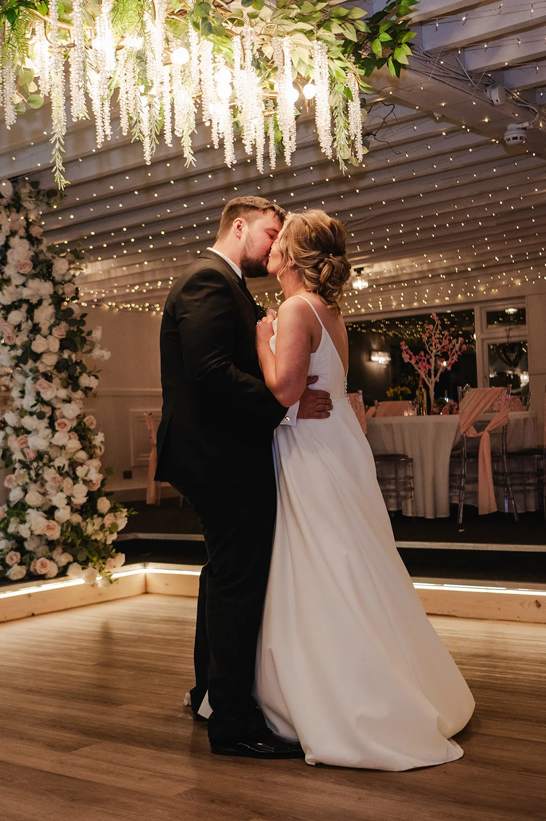 The newlywed couple share a tender kiss during their first dance under a sparkling fairy light canopy and hanging floral installation.