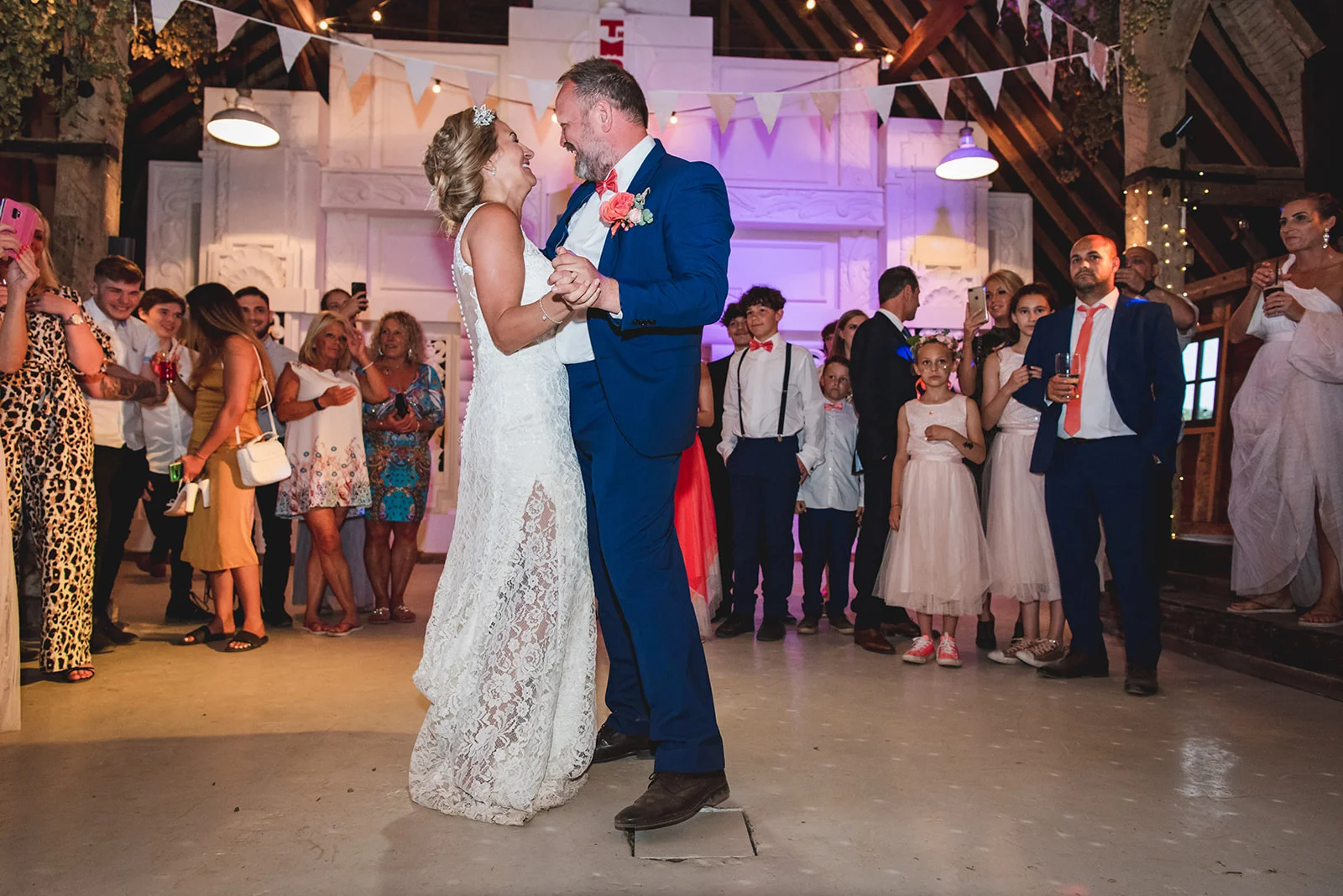 Newlyweds share a joyful first dance inside a rustic barn venue surrounded by cheering wedding guests and children.