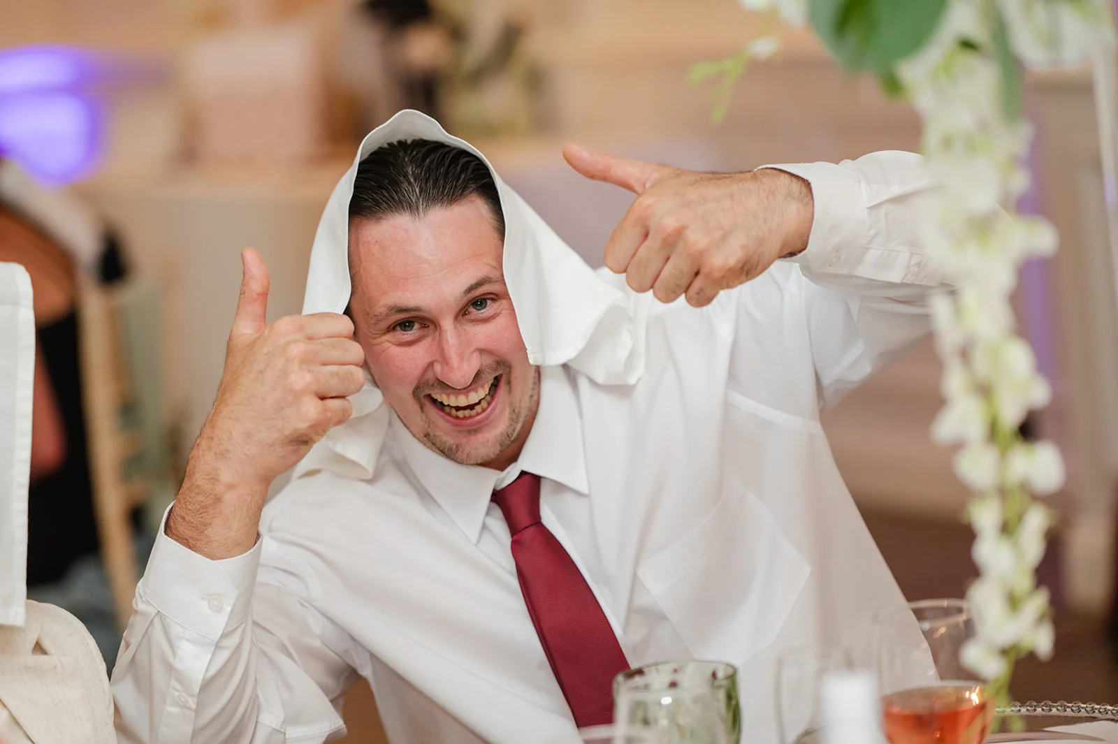 A joyful groom with a white cloth placed over his head enthusiastically gives two thumbs up while laughing at the wedding reception.