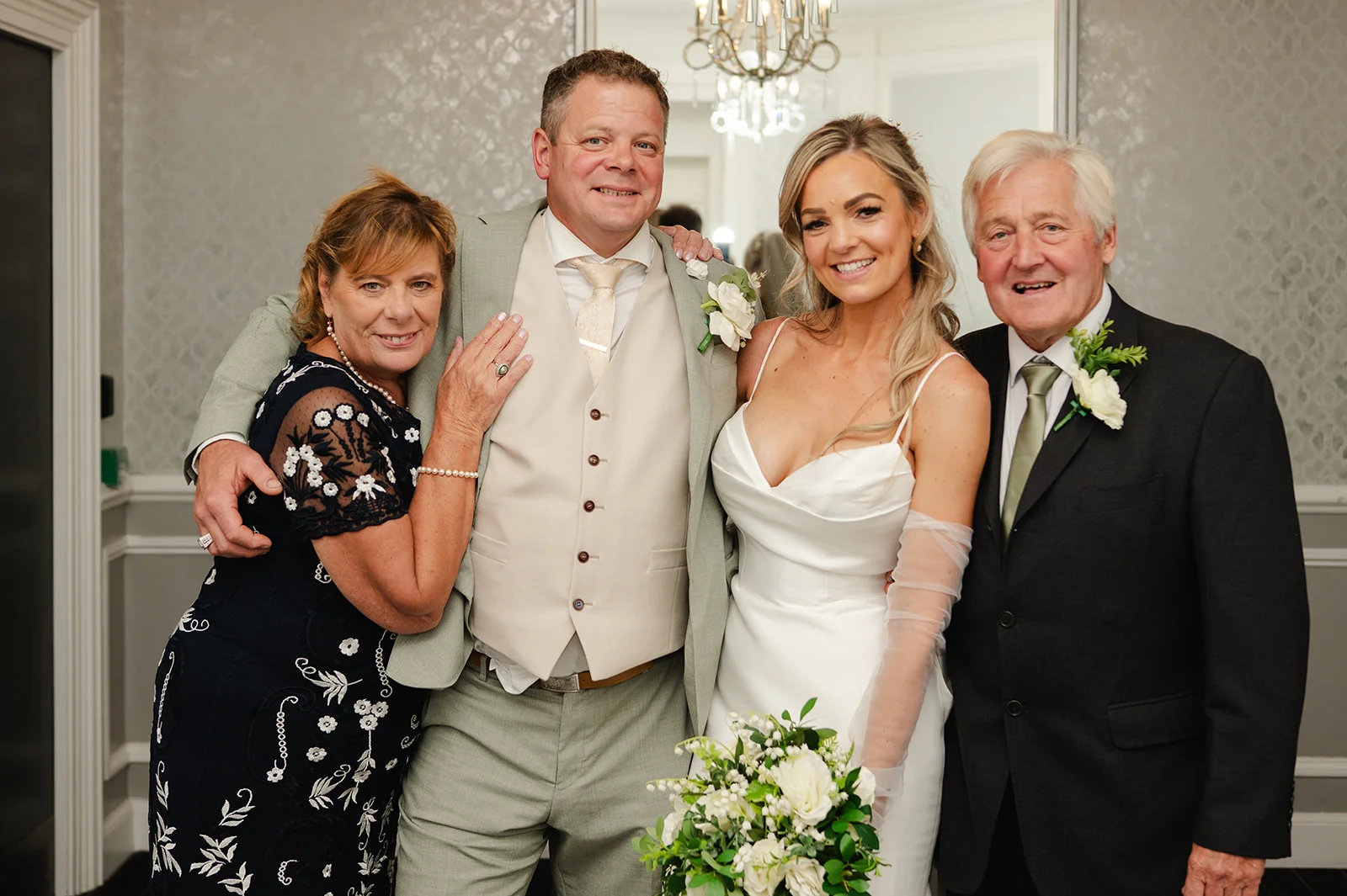 A smiling family group including the bride in a satin gown and veil gloves, the groom in a sage suit, and parents posing indoors for a formal wedding portrait.