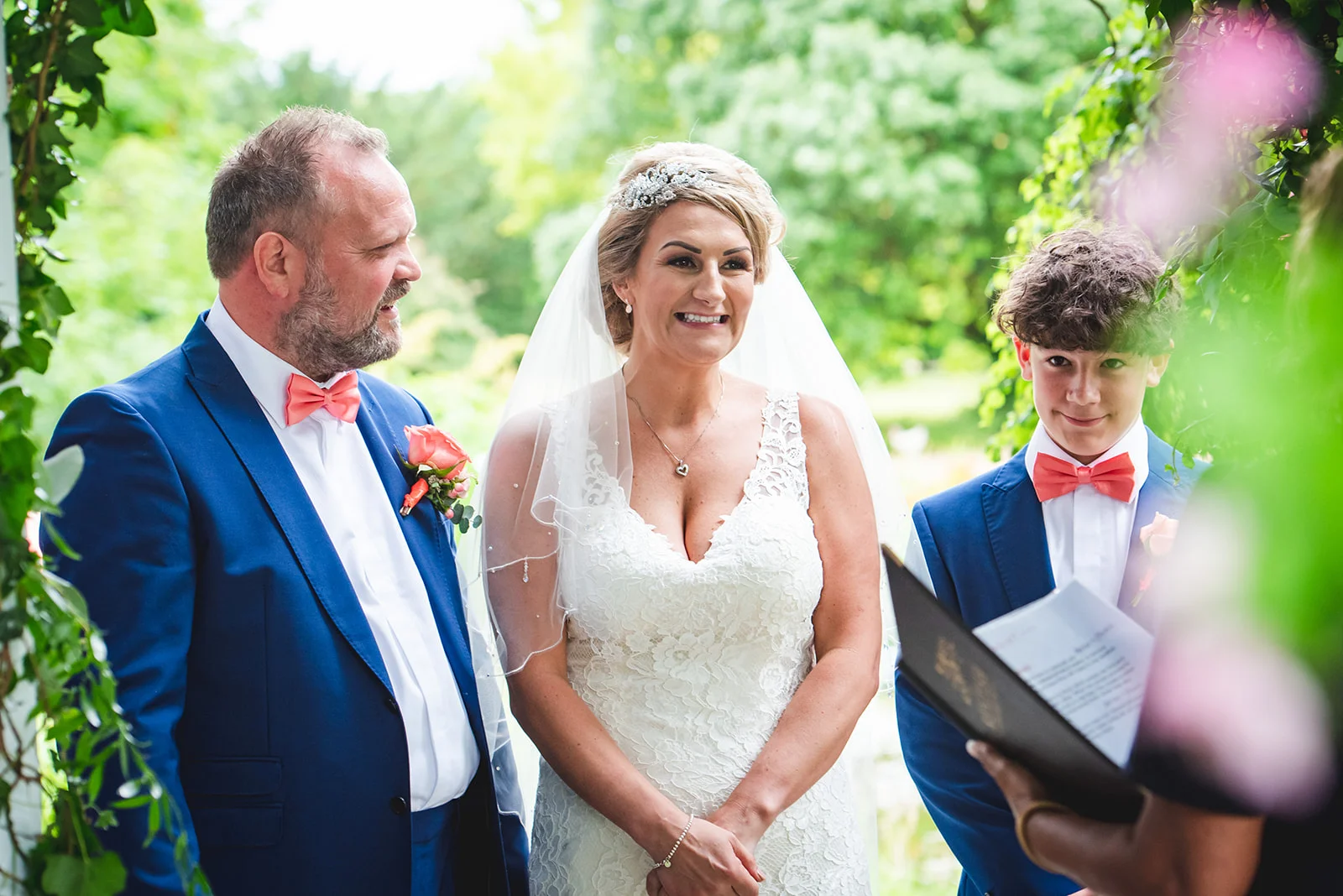 A smiling bride and groom stand together during an outdoor wedding ceremony, flanked by a young boy holding an order of service book.