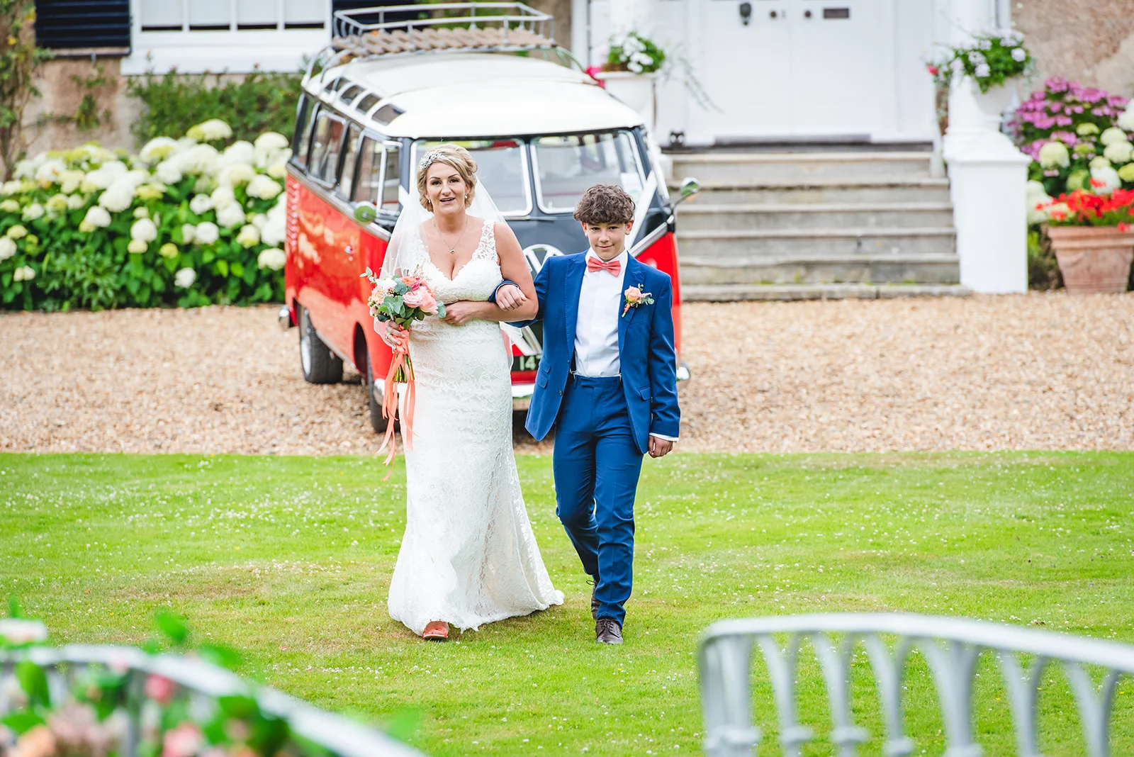 A bride in a lace gown and a young page boy walk arm in arm across a lawn in front of a vintage red and white campervan.