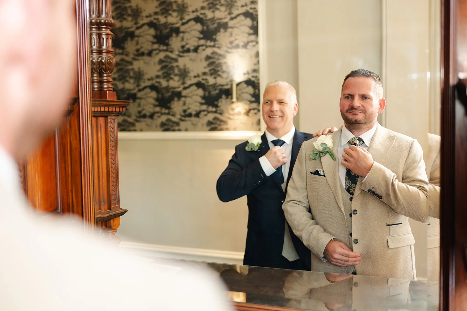 A groom in a beige suit adjusts his tie in front of a mirror while a smiling best man in a navy suit stands behind him during wedding morning preparations.